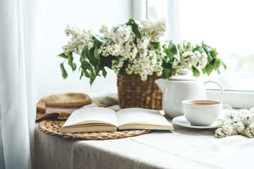 Open book, cup of tea and teapot, white lilac basket and hat, summer composition