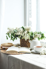 Open book, cup of tea and teapot, white lilac basket and hat, summer composition