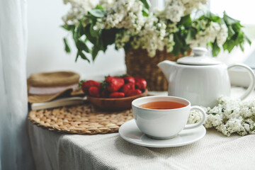 Cup of tea and teapot on windowsill and basket with white lilacs, good morning concept