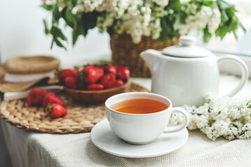Fototapeta na wymiar Cup of tea and teapot on windowsill and basket with white lilacs, good morning concept