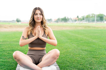 Young caucasian slim fit blond woman sitting in lotus yoga pose on fitness mat in stylish sportwear with hands as in prayer, after workout on green grass meadow near trees in summer park at daytime