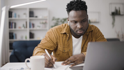 Concentrated African American man typing on laptop and taking notes in notepad, freelance