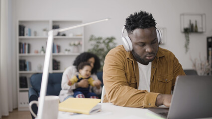 African American man working on laptop in headphones while mother is trying to comfort crying baby