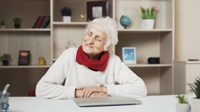 An elderly woman sits at a table in a beautiful, well-lit room. She is telling a story, smiling, and gesturing enthusiastically. She is dressed in a white sweater with a red scarf.