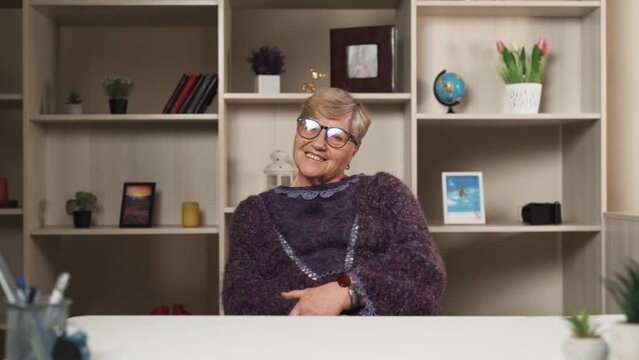 An elderly woman sits at a desk in a modern office or at home. She appears happy and smiling The woman has short blonde hair and is wearing a purple sweater. The woman is wearing glasses on her face.