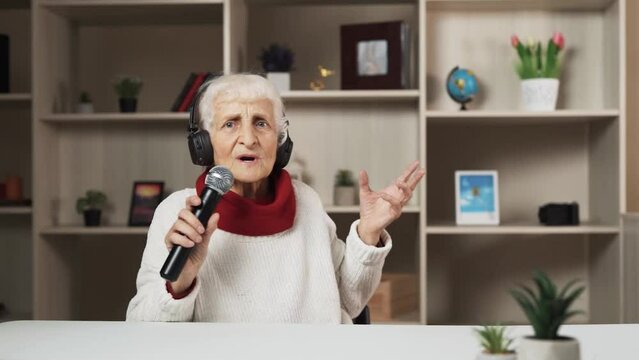 In a modern, well-lit room, there is an elderly woman sitting at a table. She has put on headphones and is holding a microphone in her hands. Additionally, she engages in dancing while seated.