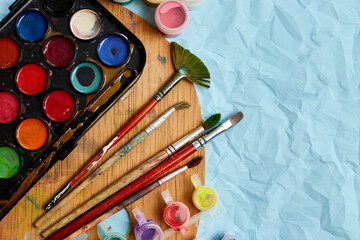 Flat lay of colorful paints, painting palette and brushes on blue background, art, painting and hobby concept, top view, copy space..