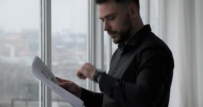 Serious and busy Caucasian businessman in a black suit stands by a window checking documents and glancing at his wristwatch. He appears to be waiting for a business meeting.
