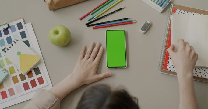 Top view of young woman touching sketchbook and looking at smartphone with chroma key green screen at desk. Stationery and modern technology concept.