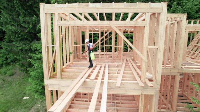 Laborer constructing wooden frame house near forest. Man treating woods, applying fire retardant using sprayer, while dressed in protective suit, helmet. Concept of modern eco-friendly construction.