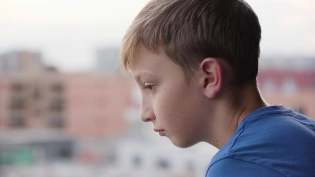 A cute young boy stands on a balcony and thinks about something - face closeup from the side - buildings in the blurry background