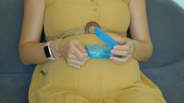 In this slow-motion video, a pregnant woman is seen sitting on a couch. She takes a pill of vitamins or food supplements from a plastic box with sections. The camera is focused on her hands and the