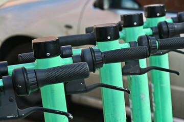 Close-up of the handlebars of a shared electric scooters parked on the street with a white car in...