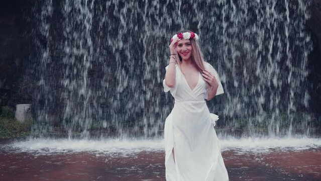 Attractive woman in white dress with a cute, angelic face poses on a background of a waterfall. Hair fluttering in the wind. Pretty lovely smile, positive emotions. Summer close-up portrait.