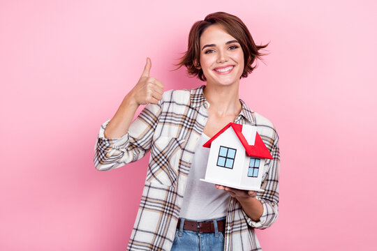 Portrait of charming positive lady hold little house demonstrate thumb up isolated on pink color background