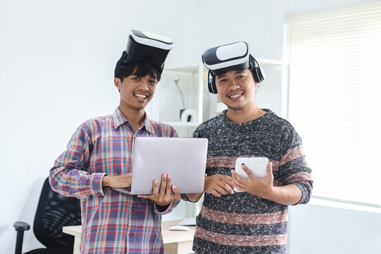 Two young millennials in virtual reality glasses holding laptop and tablet while looking into camera smiling. Teen gamers in VR helmets after win the game.