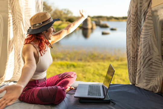 woman working with a laptop in a camper van in the middle of nature near a lake and lawn, arms open, wearing a hat, at sunset, digital nomad, having a meeting.