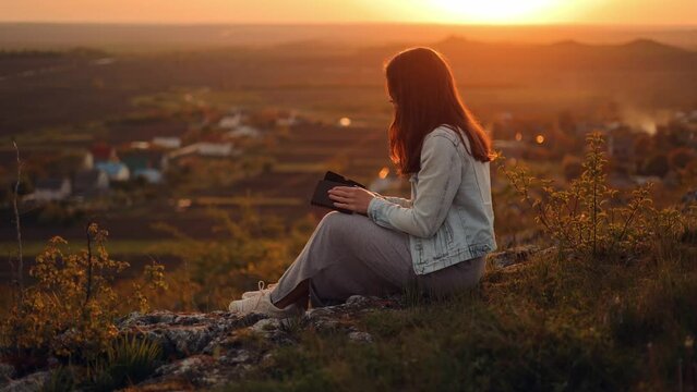 A woman reads a book in the sun. A girl is reading the Bible in the open air, holding the Bible in her hands and studying the word of God at sunset on the top of the mountain. Self-educational concep