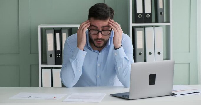 Caucasian Businessman sitting at his desk, wearing a shirt and glasses, looking through documents and becoming visibly upset. He throws the papers on the table and holds his head in frustration