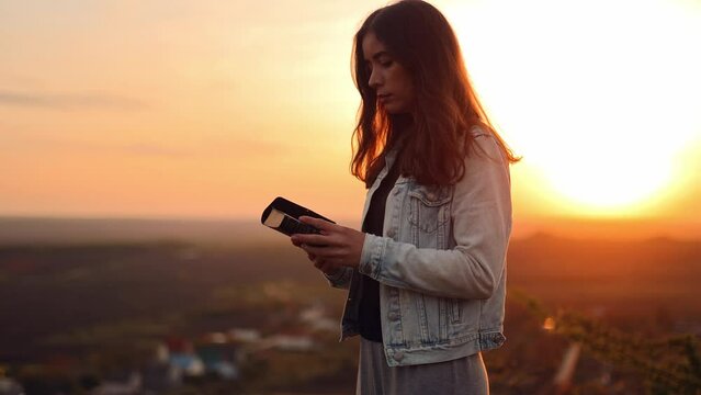 Beautiful young woman reading the bible while standing on top of a hill with a beautiful view at sunset, admiring the view and nature, relaxing alone with thoughts, slow motion
