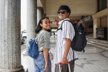 Young Asian couple smile and look back on camera. Ready to travel or have trip. Vacation mood. Carry backpack, purse or suitcase. Travel, holiday and relationship concept. 