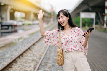 Asian young woman traveler with weaving basket using a mobile phone beside railway train station in Bangkok. Journey trip lifestyle, world travel explorer or Asia summer tourism concept.