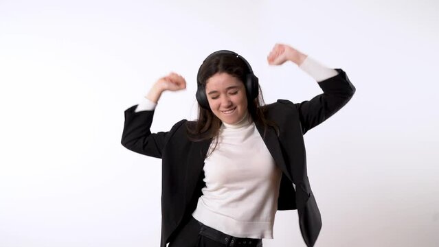 Smartly dressed young brunette against white background wearing headphones happily dancing ento music while energetically moving her hips