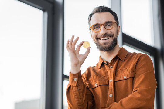 KYIV, UKRAINE - OCTOBER 18, 2022: bearded businessman in fashionable shirt and eyeglasses holding golden bitcoin and smiling at camera near blurred windows in modern office on blurred background