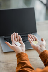 KYIV, UKRAINE - OCTOBER 18, 2022: cropped view of accomplished entrepreneur holding golden and silver bitcoins on open palms near laptop with blank screen on desk in office
