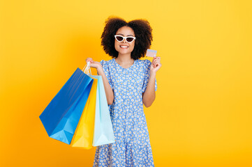 Photo of charming smiling positive african american or brazilian young woman wearing blue sundress, with sunglasses, holding paper shopping bags and credit card, stand on isolated yellow background