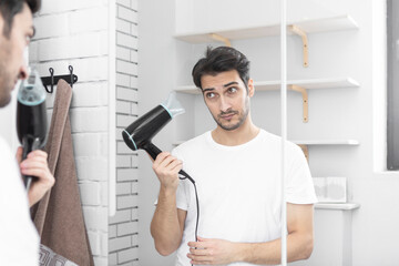 Young handsome man standing in front of the mirror in the bathroom and holding hair dryer. Morning routine and making hair style concept