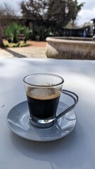 Small glass cup of black coffee with a metal handle in a white plate on the terrace