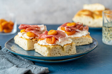 Focaccia sandwiches with prosciutto, soft cheese stracciatella, dry apricots on a blue plate, ingredients, fresh drink backside over dark blue and grey background