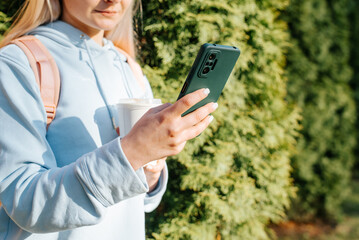 Close-up of caucasian woman in casual clothes using smartphone outdoors. Woman holding mobile phone and cup of coffee in nature