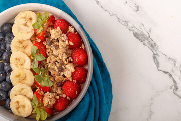 Bowl of oat granola with yogurt, fresh raspberries, blueberries, strawberries, banana and nuts on white background for healthy breakfast. Top view. Copy space. Close up