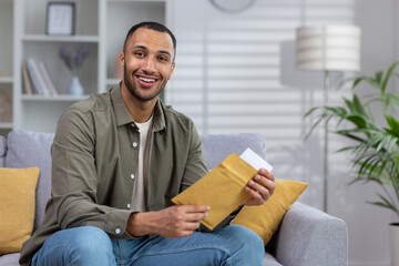 Portrait of a young Muslim man smiling looking at the camera and holding a letter, documents in an envelope delivered to his home