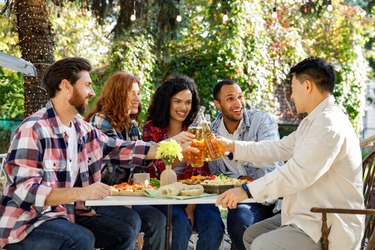 A cheerful multinational company is having fun on a picnic. Multisex friends sitting at the table outdoors celebrating eating and drinking drinks.