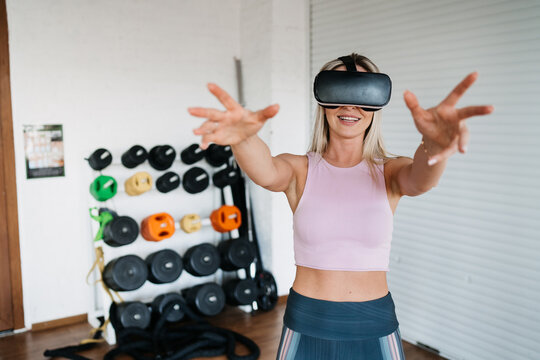 Blonde caucasian young woman in sportswear using virtual reality headset warms up using vr program for training. Cheerful american girl at exercise at sport club.