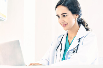 Beautiful and professional female doctor using laptop computer, working at her desk in the doctor's office.