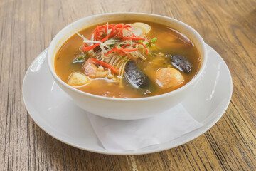 Tom Yum in white bowl isolated on wooden table with beautiful garnish. Tom Yum is a type of hot and sour Thai soup, usually cooked with shrimp or prawn with lots of herbs and spices.