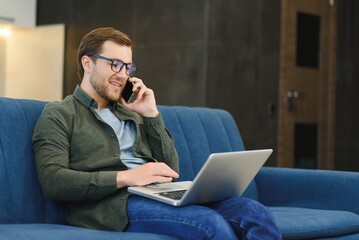 Portrait of an attractive serious young bearded man wearing casual clothes sitting on a couch at the living room, talking on mobile phone while using laptop computer.