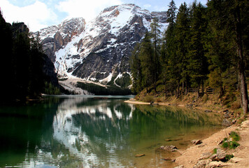 Landscape view of the emerald smooth surface of Lago di Braies lake and snow capped mountains in the background against a blue sky with clouds in the Dolomites in northern Italy