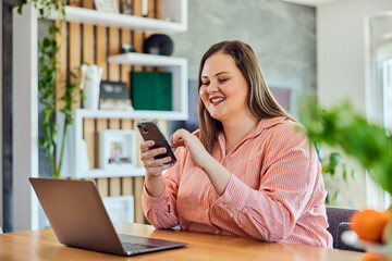 A smiling curvy businesswoman using a mobile phone while working on a laptop.