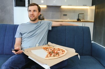 Eats pizza while watching TV show. Man with beard indoors.