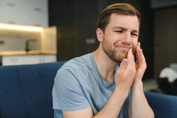 Toothache. Unhappy upset caucasian sad man sits on the couch at home, holds his hand near his...