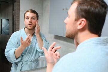 Handsome man looking at mirror and applying moisturizing cream on cheeks in bathroom. Groomed young guy doing skincare morning routine after taking a shower.