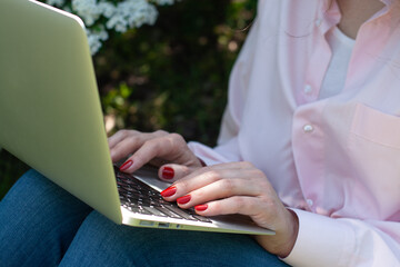 A girl in a pink shirt sits in the garden on the grass with a laptop, hands with a red manicure - 604969362