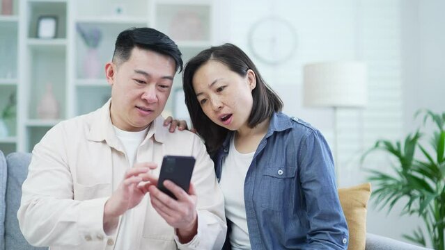 Happy asian couple using smartphone while sitting on sofa in living room at home. Smiling satisfied husband and wife browsing social networks or photos on phone, shopping online, chatting with friends