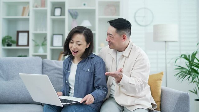 Happy asian family couple talking on video call using laptop, waving hands while sitting on sofa in living room at home. Loving wife and husband cuddles, smiles, have a conversation looking at camera
