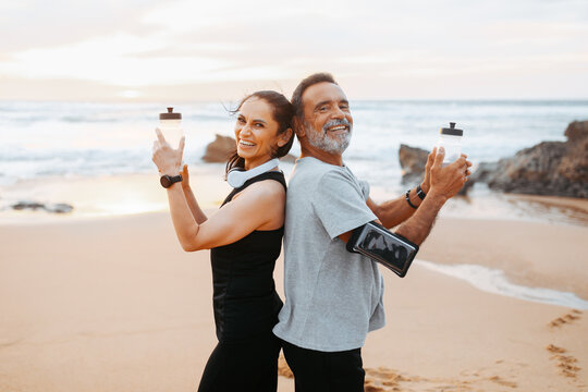 Happy mature caucasian man and woman in sportswear with phone, bottles of water have fun in morning